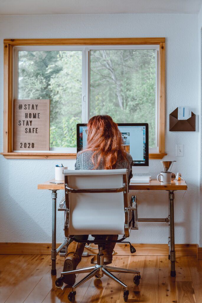 a young woman with shoulder length hair sits badly at a desk wacthing a PC screen in front of a wide window with trees outside