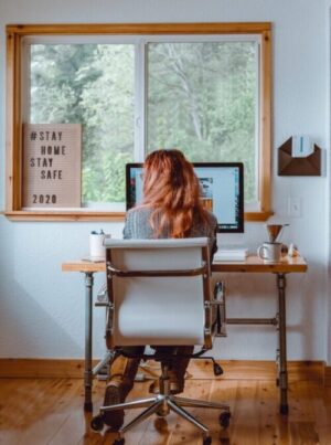 a young woman with shoulder length hair sits badly at a desk wacthing a PC screen in front of a wide window with trees outside