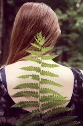 A brown-haired woman wearing a low-backed black dress has a long fern frond stretching up towards her head representing her spine.